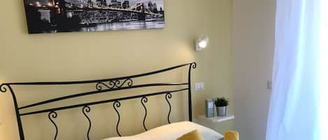 A neatly made bed with a white comforter and a decorative headboard. A small bedside table with a lamp and a potted plant is visible.