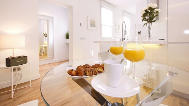 Et moderne køkken med et glasbord, lyse hvide hårde hvidevarer, og et morgenmåltid med croissanter, kaffe og appelsinjuice.