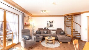 A living room with a sofa set, a round coffee table, a chair, a staircase, and a large window. - Ferienwohnung Elisabeth, 90 qm für 2-4 Personen (Reit im Winkl)