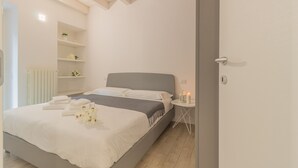 Una cama bien hecha con sábanas blancas y un cabecero gris, una mesita de noche con una vela y un pequeño estante con una planta.