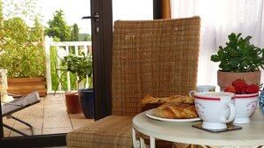 A wicker chair and table with pastries and coffee cups on a balcony with potted plants and a view of greenery.