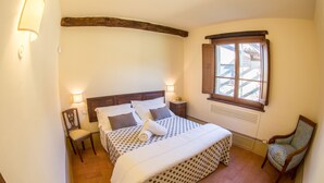 Ein Schlafzimmer mit einem Bett, zwei Nachttischen, einem Sessel, einem Fenster und einem Holzbalken an der Decke.
