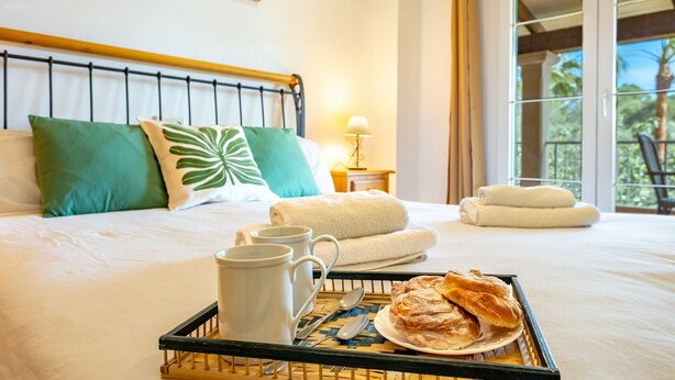 A neatly made bed with green and white pillows, a tray with a plate of pastries, two white mugs, and a spoon. - Es Recer by dracmallorca (Santanyi)