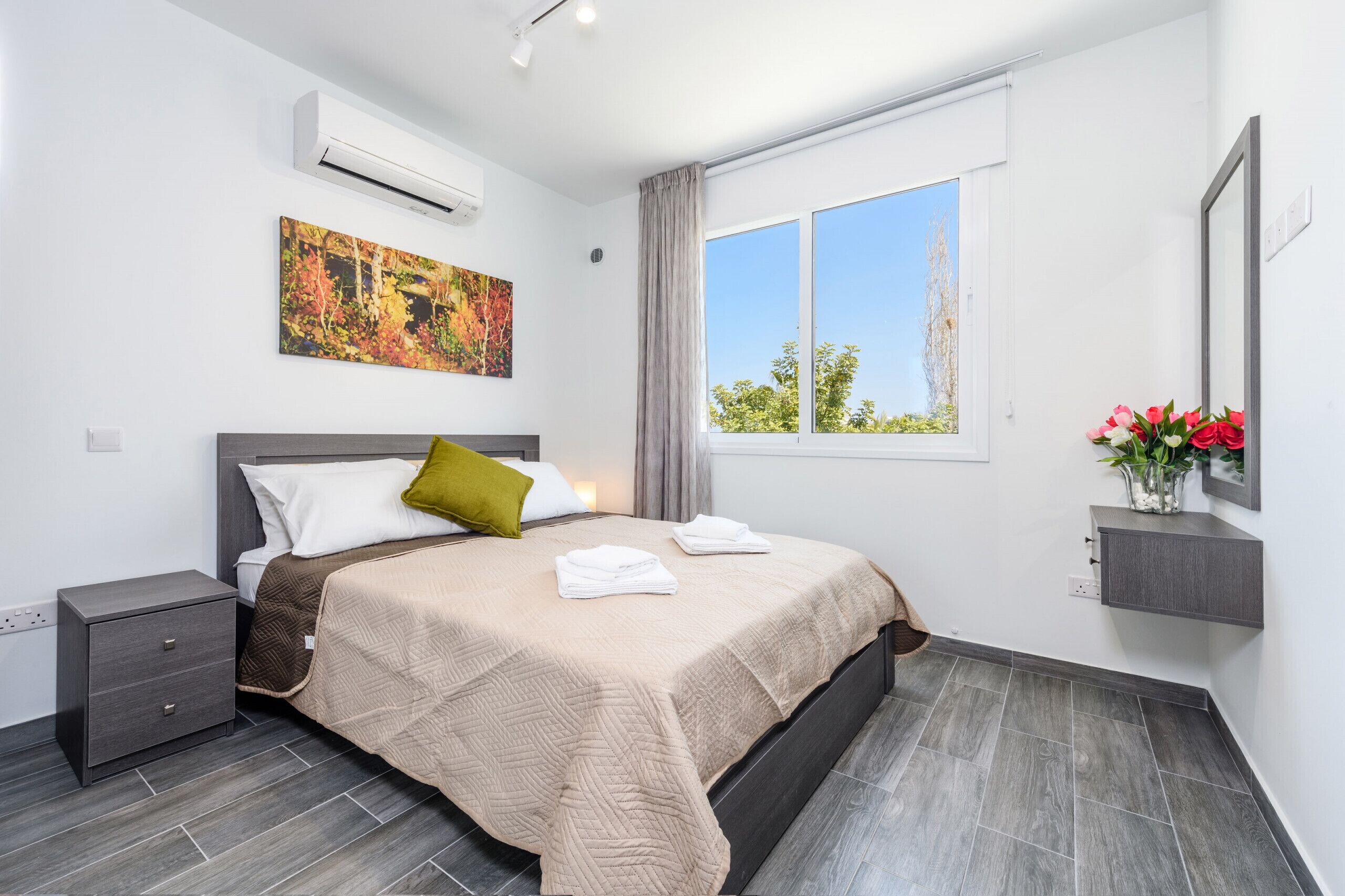 A modern hotel room with a large bed, a nightstand, a wall-mounted air conditioner, a window with curtains, and a framed picture on the wall.