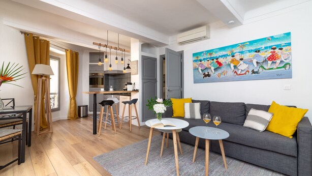 Uma sala de estar moderna com um sofá cinzento, almofadas amarelas, uma mesa redonda pequena com um livro e copos de vinho, e um quadro na parede.