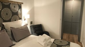 Cama bem arrumada com lençóis brancos e toalhas dobradas, uma poltrona estofada preta, uma luminária fixada na parede e um armário fechado com portas venezianas horizontais.