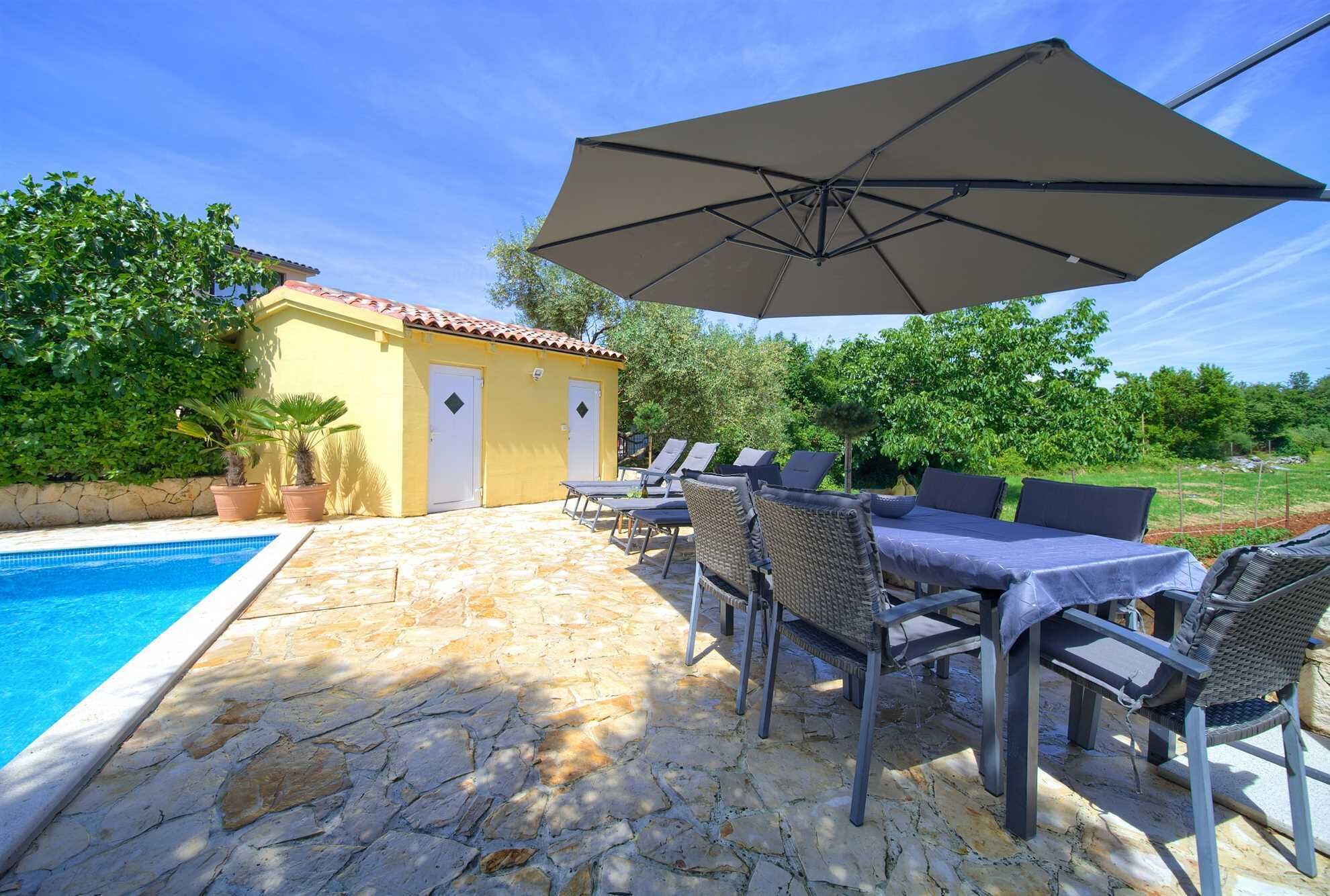 Zona de piscina con suelo de piedra, un edificio amarillo con puertas y mobiliario exterior con sombrillas.