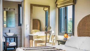 A bedroom with a large bed, a bedside table with a lamp, a dresser, a mirror, and a window with a view of greenery.