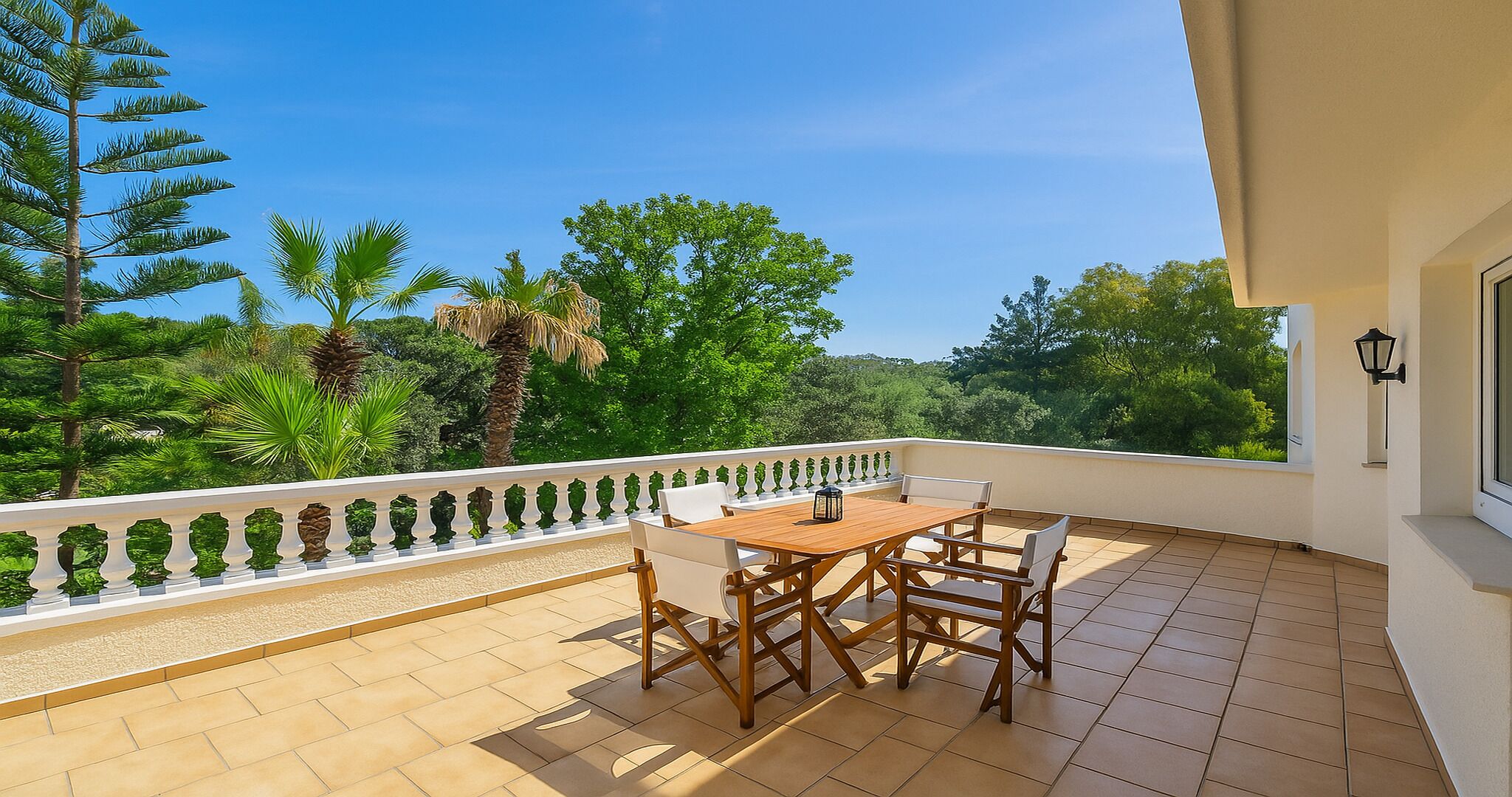 Un balcon avec une table et des chaises en bois, une balustrade blanche et une vue sur des arbres verdoyants et un ciel dégagé.