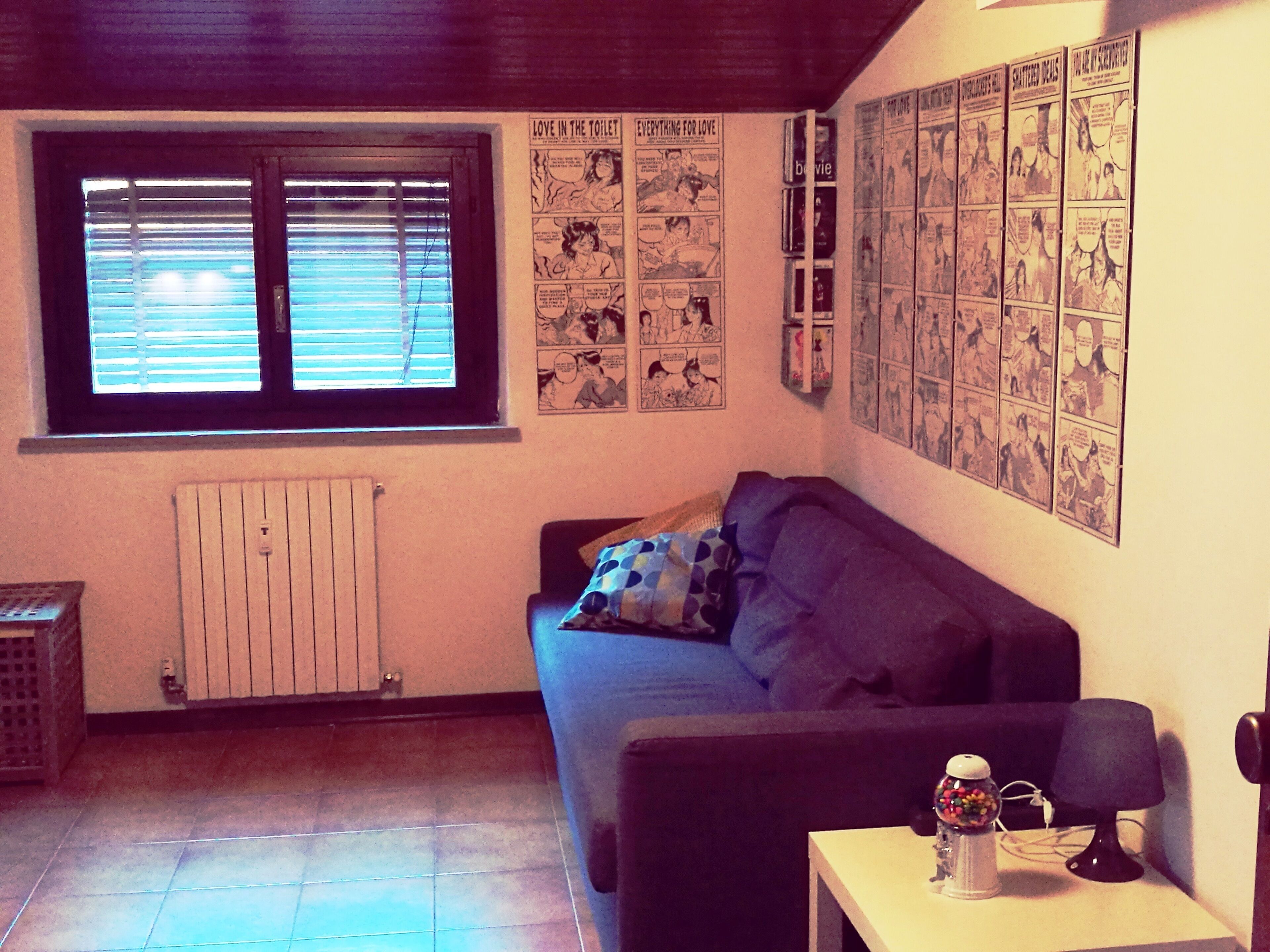 A living room with a window, a radiator, a couch, and a wall covered with comic strips.