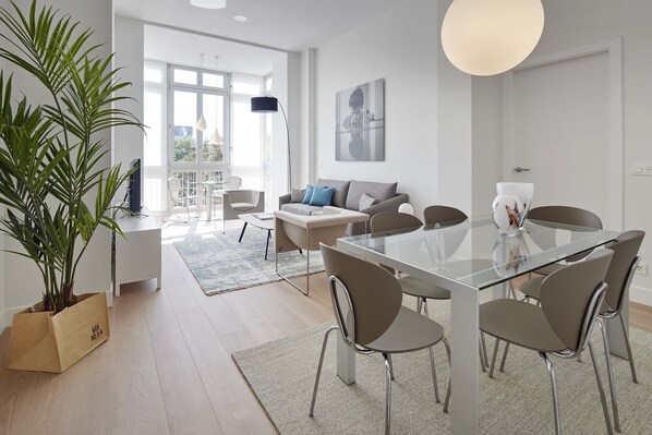 A modern living room with a glass dining table, grey sofa, and a large potted plant.