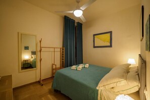 A hotel room with a bed, a standing fan, a mirror, a framed picture, and a window with curtains.