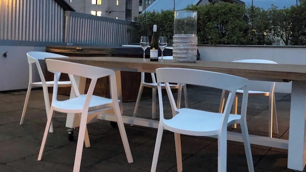 Un área de estar al aire libre con sillas de plástico blanco y una mesa de madera, que incluye un florero de vidrio y copas de vino.