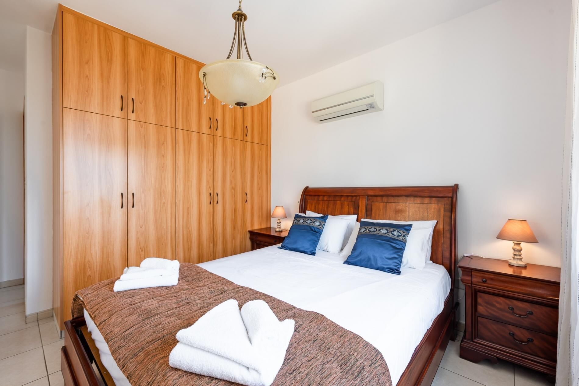 A bedroom with a wooden wardrobe, a bed with white and blue bedding, and two bedside tables with lamps.