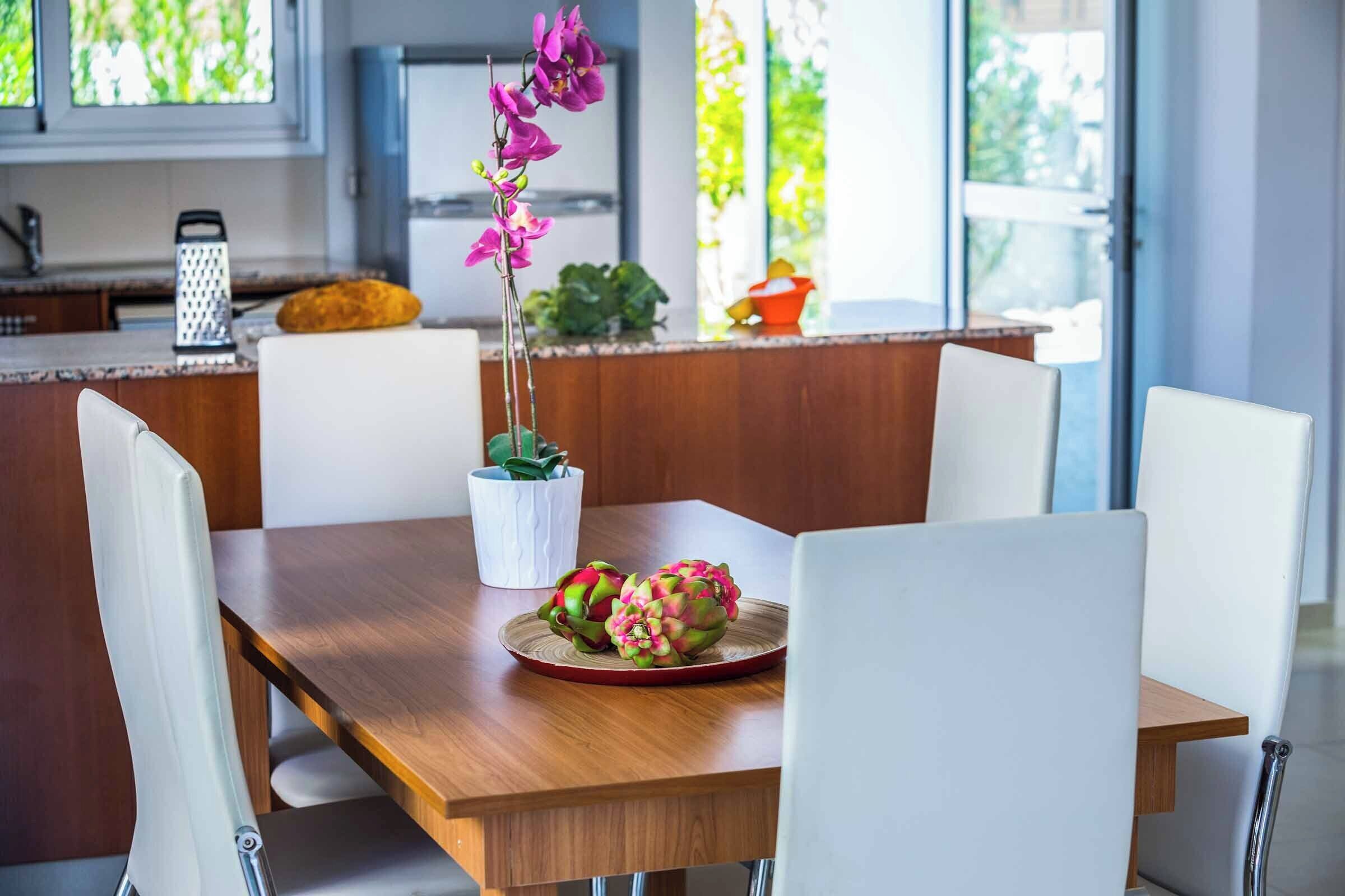 A modern dining area with a wooden table, white chairs, a potted orchid, and dragon fruit on a red plate.