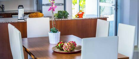 A modern dining area with a wooden table, white chairs, a potted orchid, and dragon fruit on a red plate.