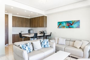 Een moderne woonkamer met een beige bank, een bijzettafeltje en een eethoek, met een keuken op de achtergrond.