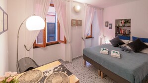 A bedroom with a bed, bedside table, lamp, curtains, window, and a bookshelf.