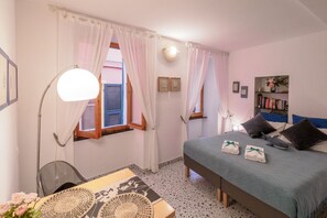 Un dormitorio con cama, mesita de noche, lámpara, cortinas, ventana y estantería.