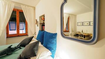 Een slaapkamer met een bed, kussens, een nachtkastje, een spiegel en een raam met gordijnen.
