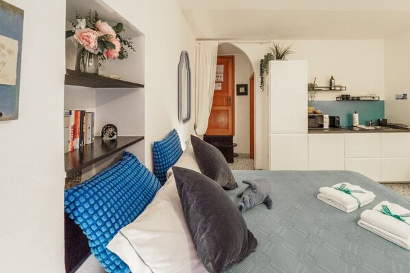 A bedroom with a bed, pillows, a bedside table, a mirror, a shelf with books, a vase with flowers, and a view into the kitchen.