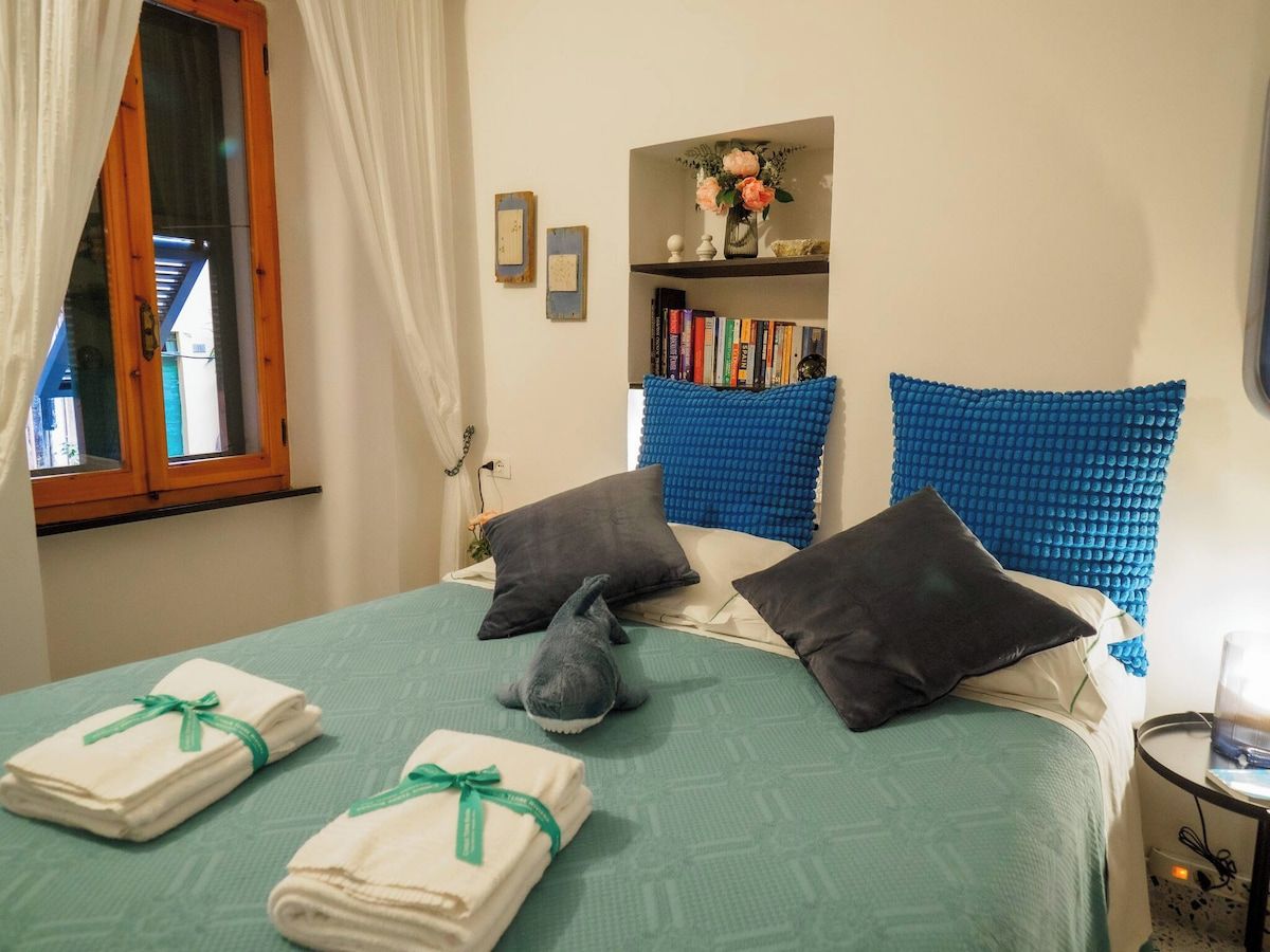 A hotel room with a bed, pillows, a bookshelf, and a window with curtains.