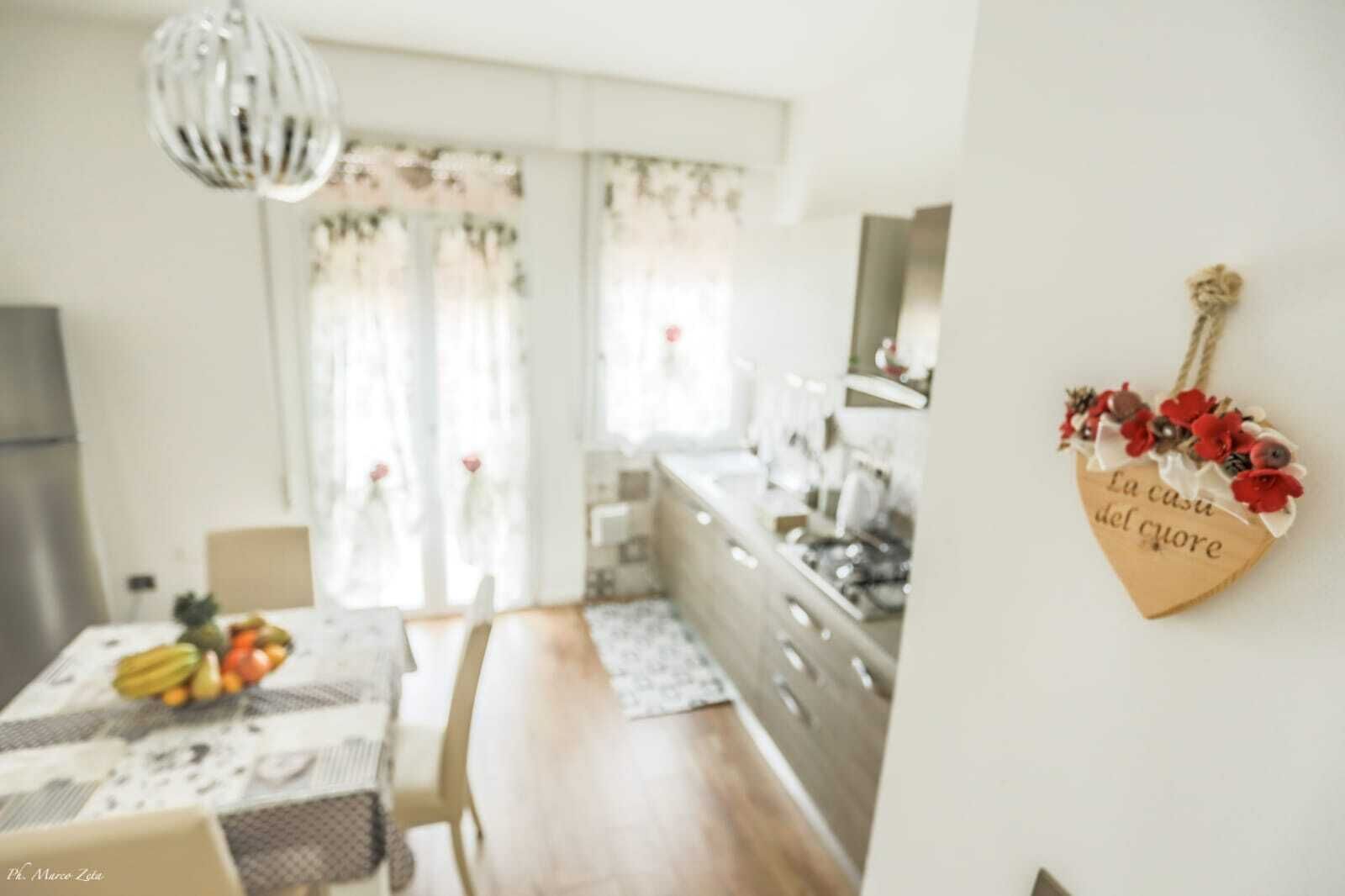 A kitchen with a dining table set for two, a fruit bowl, and a heart-shaped decoration on the wall.