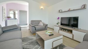 Ein modernes Wohnzimmer mit einem grauen Sofa, einem Couchtisch mit Fernbedienung, einem Fernseher auf einem Holzboden und einer kleinen Pflanze.