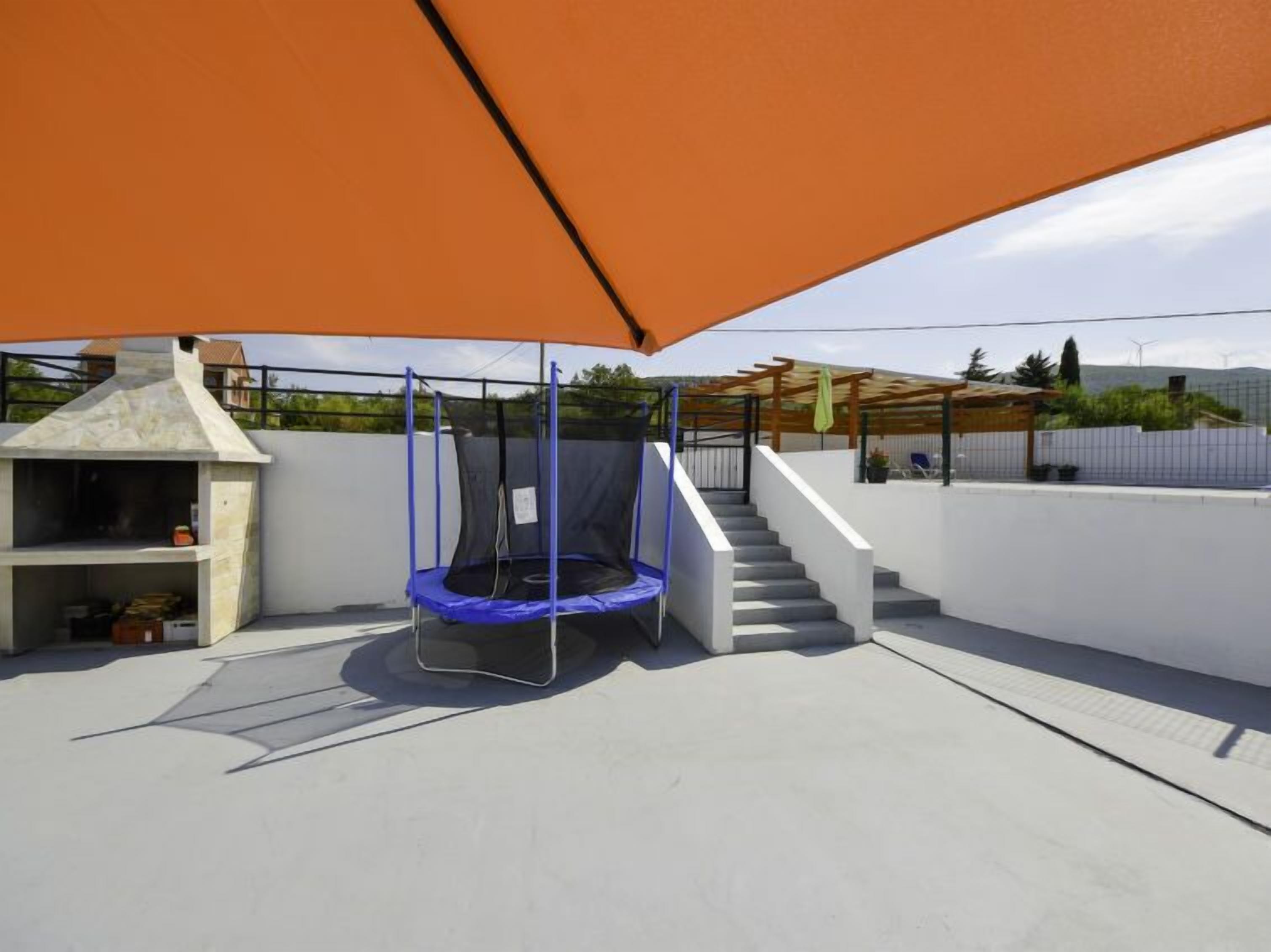 Un patio en la azotea con un trampolín, una parrilla para barbacoa y una pérgola con un parasol verde.