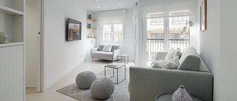 A modern living room with a grey sofa, a coffee table, and a grey rug. There is a window with a view of buildings outside.