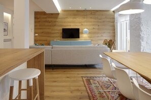 A modern living room with a wooden accent wall, a flat-screen TV, a sofa, a dining table with chairs, and a patterned rug.