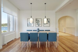 A dining area with a wooden table, blue chairs, and pendant lights. There are framed artworks on the wall and a view of the outdoors through large windows.