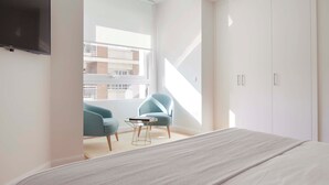 A modern bedroom with a bed, a TV mounted on the wall, a window with a view of the outside, and a chair with a table.