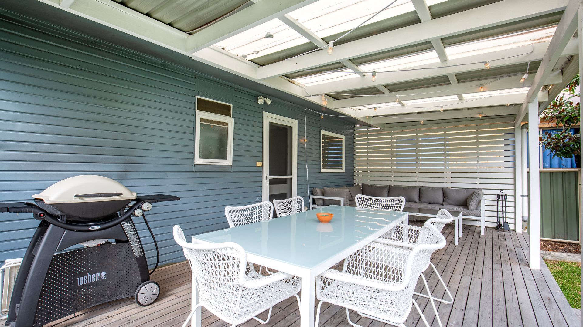 A covered patio with a grill, white wicker chairs, a glass table, and a grey sofa.