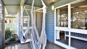 A hammock hanging from a porch railing, a window with a patterned curtain, and a view of a house in the background. - Hayes @ Jervis Bay - Pet Friendly Award Winner - 4 Mins to Beach (Callala Beach)