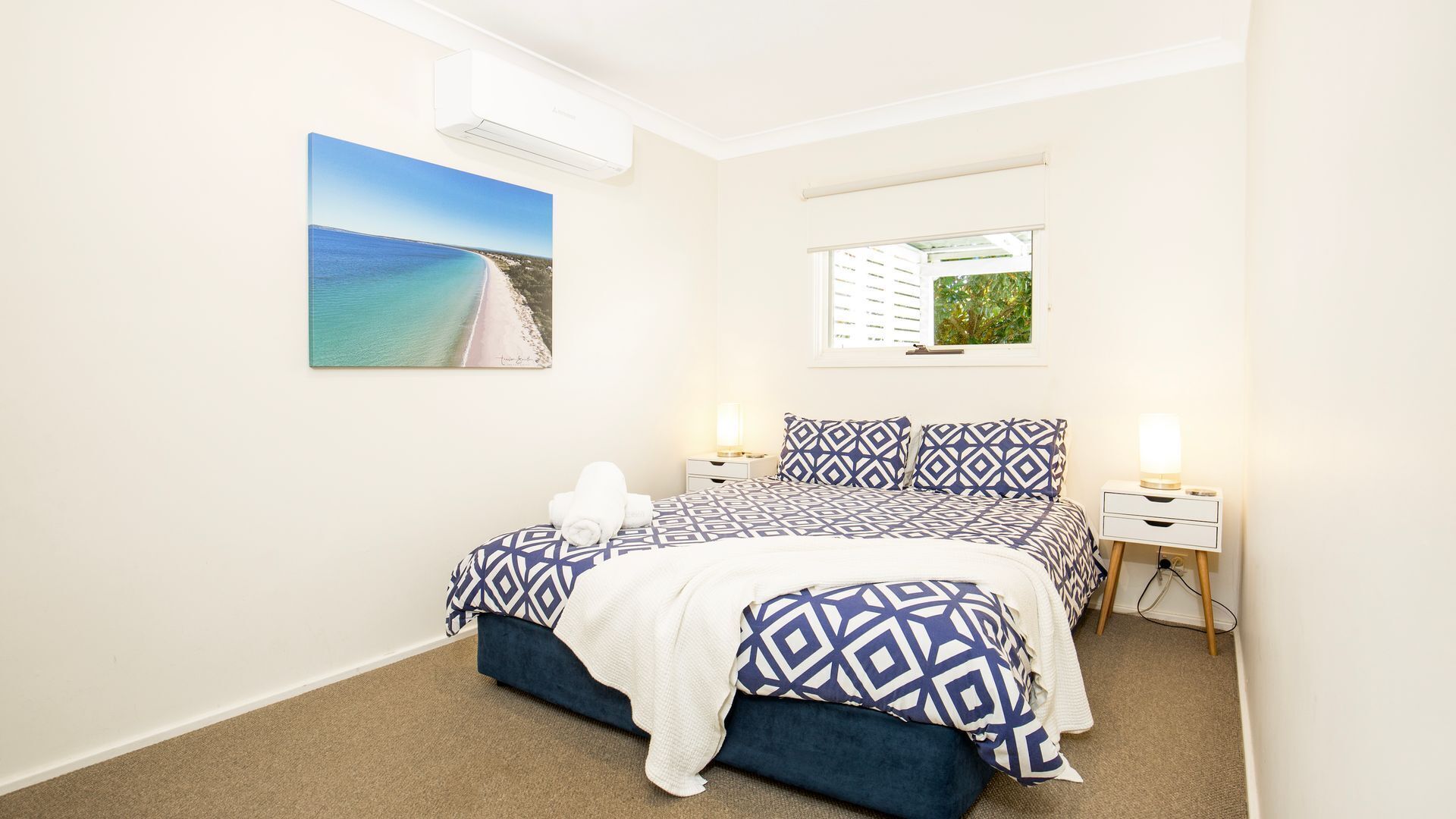 Une chambre à coucher comprenant un lit, deux tables de chevet, une commode, une fenêtre avec stores et un climatiseur fixé au mur.