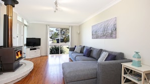A living room with a wood-burning stove, a grey sofa, a television, and a painting on the wall. - Hayes @ Jervis Bay - Pet Friendly Award Winner - 4 Mins to Beach (Callala Beach)