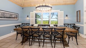 Une salle à manger avec une grande table en bois entourée de chaises, un lustre et une vue sur la verdure grâce à de grandes fenêtres.