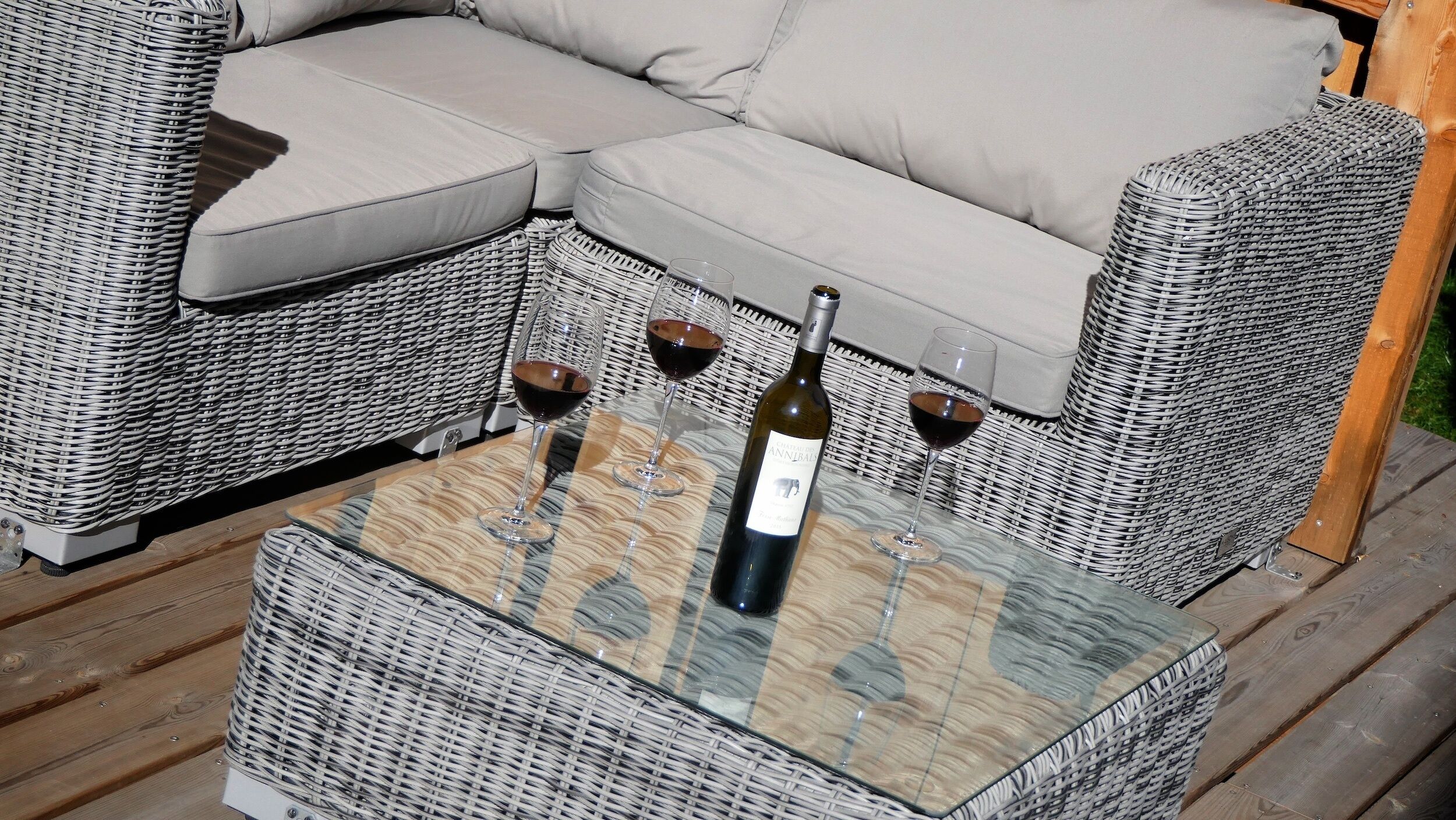 Een rieten bank met een kussen, een glazen tafel met een fles en glazen, en een geweven stoel.