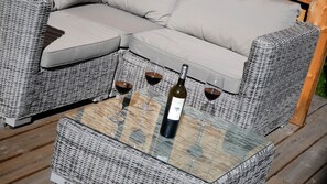 Un sofá de mimbre con una mesita de vidrio al frente, tres copas de vino y una botella.