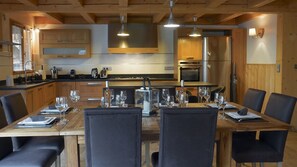 Un comedor con una mesa de madera dispuesta para seis personas, rodeada de sillas. Se ve una cocina con armarios de madera y electrodomésticos de acero inoxidable al fondo.
