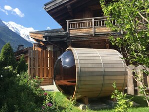Une cabane en bois avec une grande structure en forme de tonneau dans le jardin, entourée d’une végétation luxuriante et dominée par une chaîne de montagnes en arrière-plan.