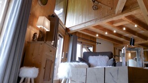 L’intérieur d’un chalet en bois, avec un plafond haut, un porte-skis, un canapé avec des coussins blancs et gris, et une petite table avec une lampe.