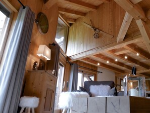 L’intérieur d’un chalet en bois, avec un plafond haut, un porte-skis, un canapé avec des coussins blancs et gris, et une petite table avec une lampe.