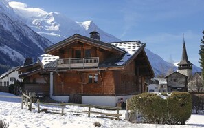 A wooden chalet with a balcony, set against a backdrop of snow-covered mountains and a church. - 5* CHALET WITH SAUNA - LES 3 CIMES BLANCHES (Chamonix)