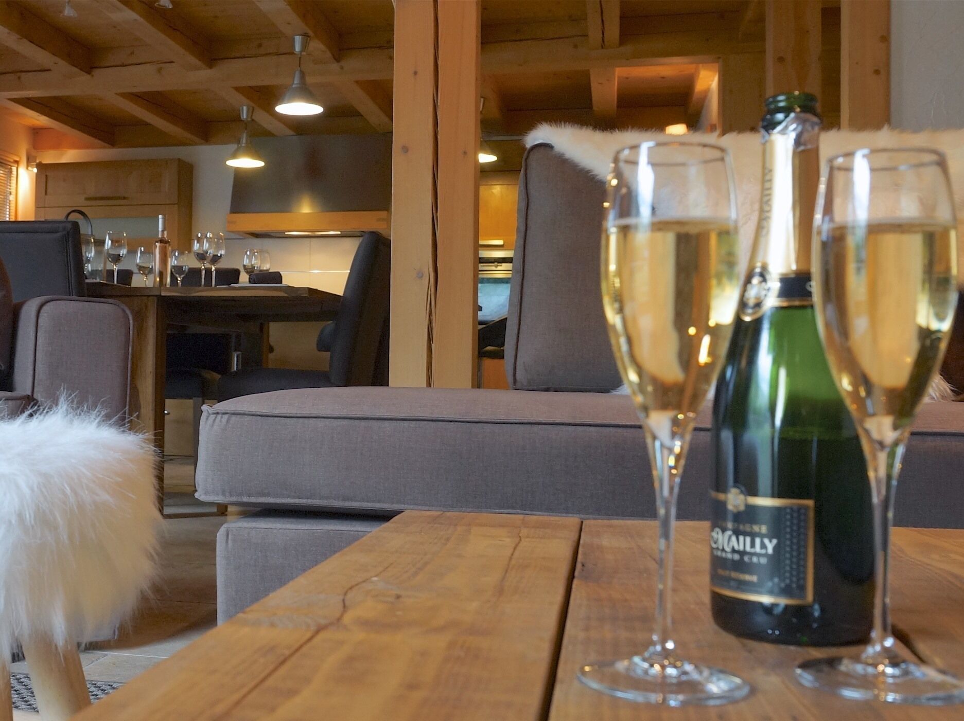 Une pièce chaleureuse avec des poutres apparentes, un coin repas avec des chaises et une table, et une bouteille de vin posée sur la table.