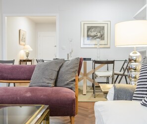 Ein Wohnzimmer mit einem violetten Sofa, einem weißen Sessel und einem Glastisch. Es gibt eine Essecke mit Tisch und Stühlen, und eine Lampe auf einem Beistelltisch.