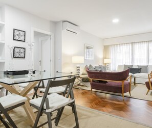 Ein modernes Wohnzimmer mit einem Glastisch, zwei Sesseln, einem Sofa und einem Couchtisch. An den Wänden befinden sich gerahmte Bilder, eine Lampe und eine Klimaanlage.
