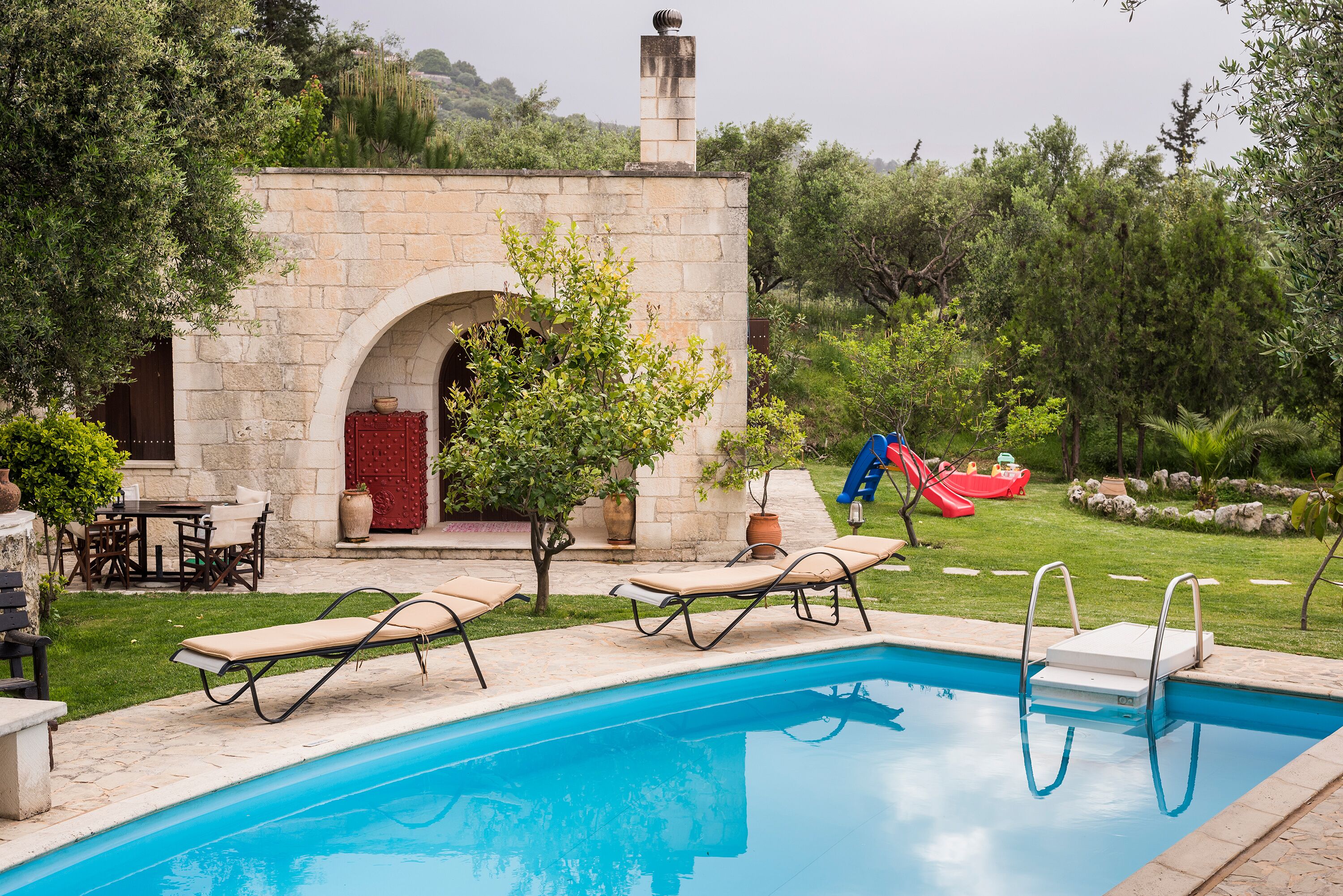 Ein Swimmingpool mit Liegestühlen, ein Steinhaus mit Bogen und eine Rutsche in einem üppigen Garten.