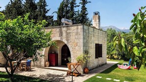 A stone building with a chimney, a small patio with a table and chairs, and a garden with trees and a swing set.